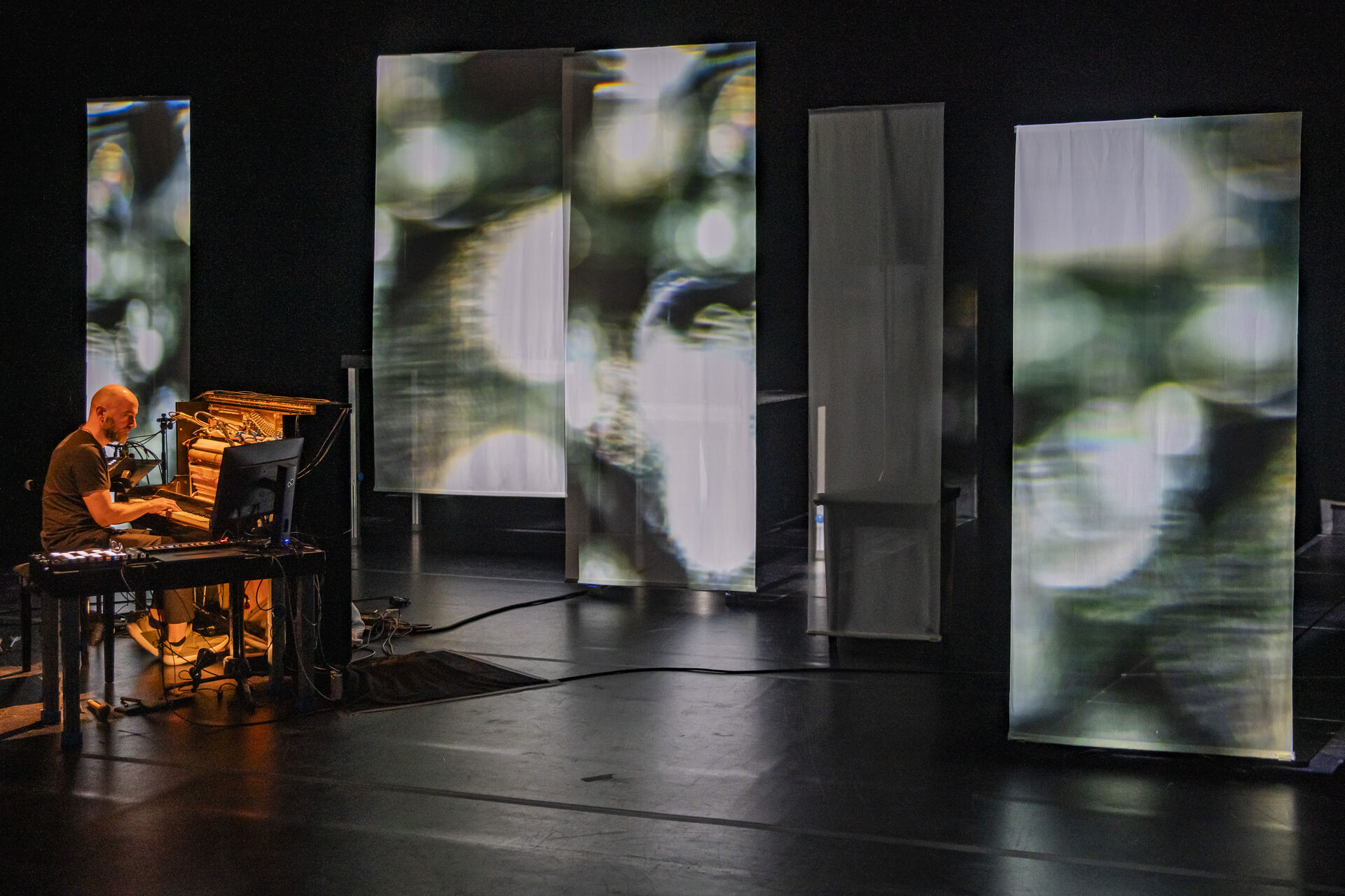 Expanded Piano II: Gasparatos at the piano on a dark stage, surrounded by tall sheer panels carrying diffuse green-and-white light projections. Photo by Andreas Simopoulos.